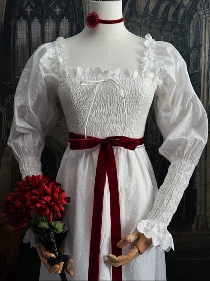 A white long-mutton-sleeve historical-style chemise dress displayed on a mannequin, with a dark red velvet ribbon sash belt at the waist, a red neck velvet choker, a small crown, and a bouquet of red flowers, set against a dark gothic interior background.